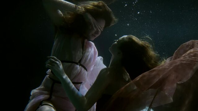 two women are floating in the dark water, looking and touching each other