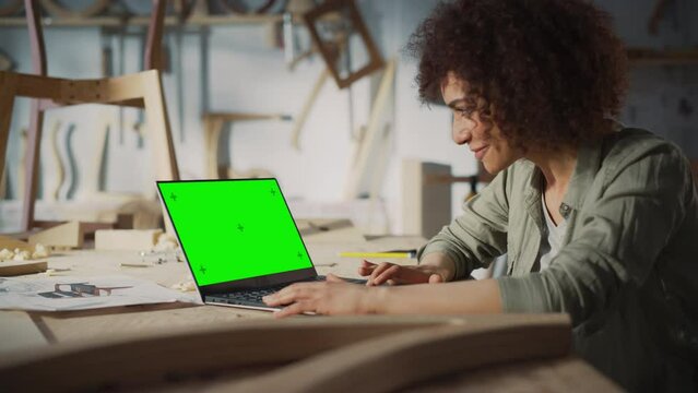 Arc Shot of a Multiethnic Furniture Designer Working on Laptop Computer with Green Screen Mock Up Display. Creative Female Preparing a Design for Carpentry Project.
