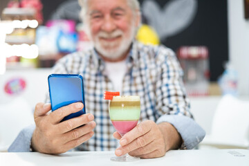 Senioro man using phone sitting close to a glass of local specialty of Canary island called barraquito, made with condensed milk, coffee and liqueur