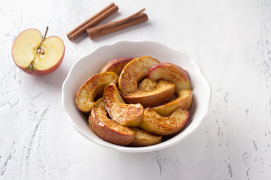 A white bowl of baked apple slices with cinnamon on a light blue background. Delicious homemade vegan dessert