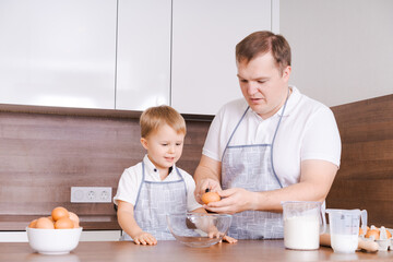 Obraz na płótnie Canvas Dad and son in kitchen, father teaches child to break eggs into a bowl. Cooking together at home. The son helps his parents. Happy father's day time