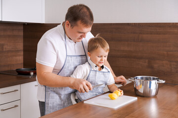Obraz na płótnie Canvas Concept food and nutrition. Shot of two cheerful dad and son posing in kitchen peeling potatoes going to cook vegetarian food for dinner together, wearing identical aprons