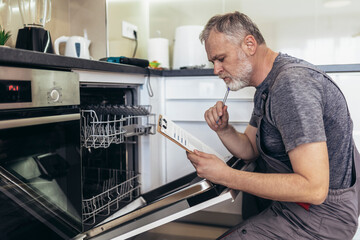 Male Technician Sitting Near Dishwasher Writing On Clipboard In Kitchen.