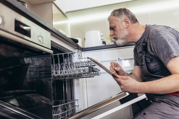 Male Technician Sitting Near Dishwasher Writing On Clipboard In Kitchen.