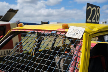 Closeup of a dented yellow stock car with number 228 and wire mess instead of a windshield on a sunny day with clouds.