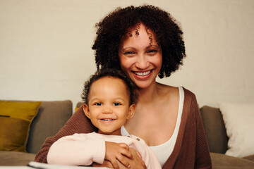 Happy biracial mother and daughter smiling and looking at camera in living room at home