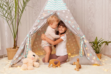 Portrait of cute little sisters playing in wigwam hugging while playing together, elder girl and toddler baby sitting in peetee tent, having fun with interest. © sementsova321