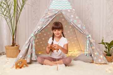 Indoor shot of charming smiling little girl with braids wearing white t shirt posing in wigwam and playing with wooden eco toy, looking at camera and expressing positive emotions. © sementsova321