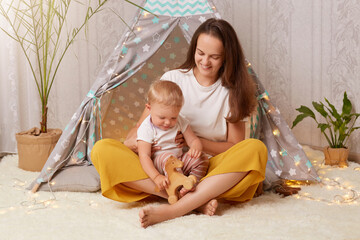 Horizontal shot of beautiful Caucasian young mother wearing white t shirt sitting in wigwam and holding her baby daughter on her knees and playing with wooden eco toy. © sementsova321