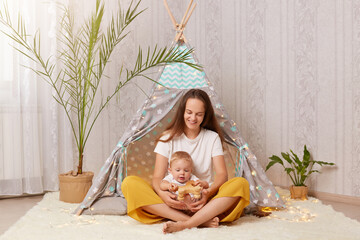 Smiling satisfied woman with long dark hair wearing white t shirt sitting in peetee tent with her baby child and holding wooden horse in hands, playing with toddler daughter at home in wigwam. © sementsova321
