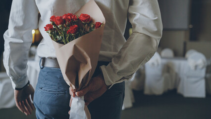 Loving man is hiding red roses in craft paper behind his back bringing beautiful bouquet for his date in restaurant. Flowers, romantic relationship and dating concept. - obrazy, fototapety, plakaty