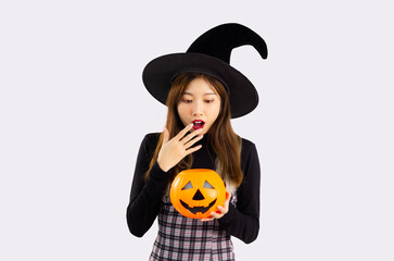 Fototapeta premium Young asian pretty woman in black halloween costume wearing witch hat and holding pumpkin lantern posing surprised and excited on white background.