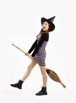 Young asian witch girl in black halloween costume wearing witch hat and holding witch broom posing on white background.