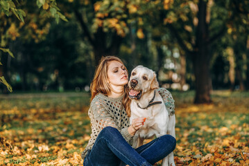 A beautiful blonde is sitting in yellow leaves in a park and playing with her labrador.