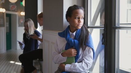 Portrait of sad African-American teen student sitting on window sill in corridor. 
