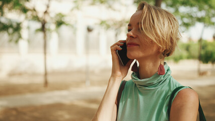 beautiful smiling woman with short blond hair in casual clothes walks through the city square, talking on cellphone