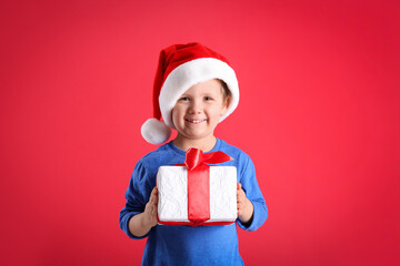 Cute little boy in Santa Claus hat holding gift box on red background