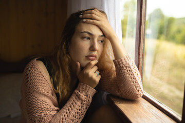 A woman sits by the window and is sad. Depressed mood. Sad woman near the window. Anxiety.