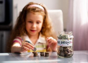 A sweet girl with full piggy bank of coins. On the piggy bank there is an inscription for dream. The girl is at home. She sits at a table and counts the coins. The money. Savings. Childhood.