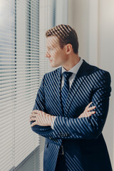 Waist up portrait of handsome male financier looks attentively in office window, has thoughtful expression, rests after conference, has work break, keeps hands crossed, wears elegant clothes