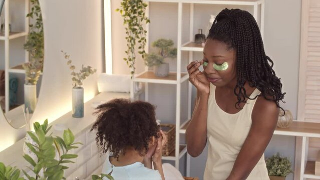 Medium slowmo of young Black woman and her cute little daughter bother wearing hydrogel undereye patches chatting in bathroom while doing morning routine