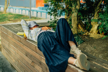 a young woman lies on a wooden bench in summer, covering her face with a baseball cap