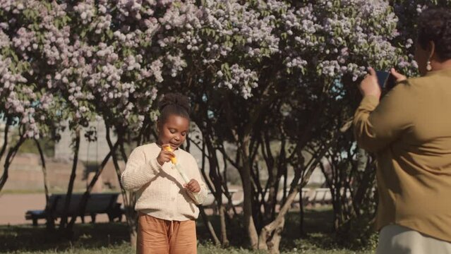 Slowmo of plus size African American woman taking pictures of her cute little daughter blowing soap bubbles in park on sunny day