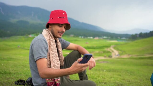 Hipster traveler in hat ralaxing in nature and using internet on smartphone, tourist boy chatting on cellphone on background of panorama horizon mountain landscape copy space.