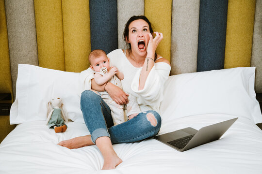 Cheerful mother using computer while stressed with infant together at home