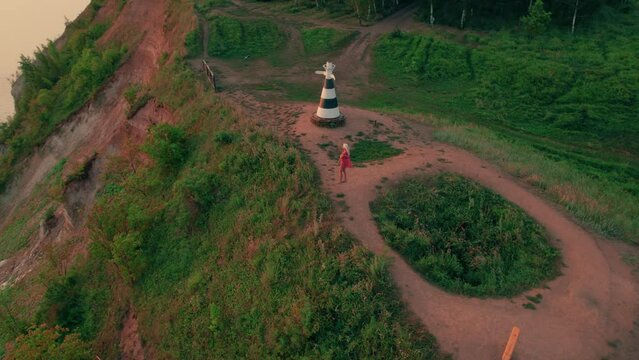 Shot on a drone. A girl in a red dress stands on a hill on the steep bank of the river and enjoys the sunrise. Enjoying the dawn.