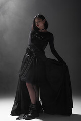 full length of asian woman with closed eyes standing in black gothic dress on dark grey with smoke.