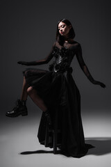 Fototapeta na wymiar full length of asian young woman in black gothic dress and gloves sitting on high chair on dark grey.