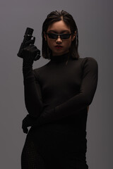 dangerous asian woman in total black outfit and stylish sunglasses holding gun isolated on grey.