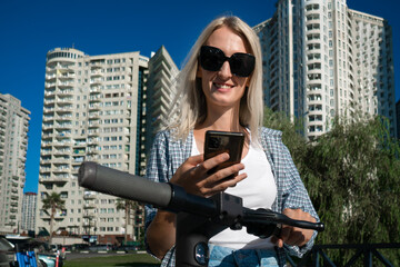 Fototapeta na wymiar Portrait of a young smiling blonde woman in sunglasses with a scooter and a mobile phone in her hands on a sunny day against the background of high-rise buildings, blue sky