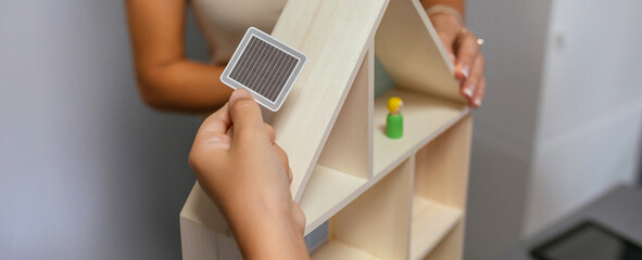Unrecognizable student placing a solar panel on the roof of a toy wooden house in an ecology classroom