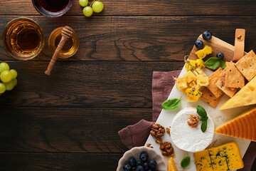 Assortment of cheese, honey, cracker, blueberries, grapes with red and white wine in glasses antipasto server on white marble board on old rustic wooden table background. Flat lay, copy space.