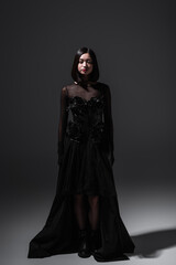 full length of asian young woman in black gothic dress and boots standing on dark grey.