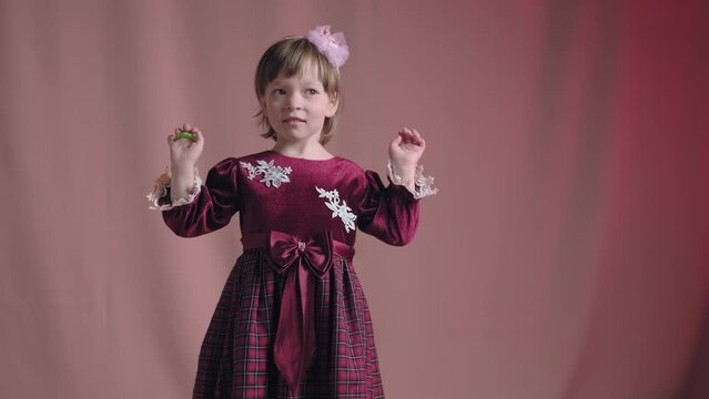 Cute little girl in an vintage burgundy dress is playing with a rattle toy. The concept of children theater productions and developing acting circles. Pink background