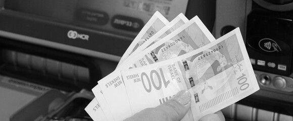 Loan Concept. Woman hand holding 100 Israeli Shekel notes on  background of Cash Machine, ATM. Black and white - obrazy, fototapety, plakaty