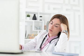 Close up portrait of tired female doctor sitting at the desktop and working on laptop in the office of modern clinic