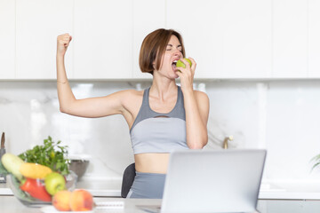 Young slender food blogger eating apple and demonstrating her biceps to her subscribers. Woman cooking healthy food using fruits and vegetables and biting apple at camera on laptop