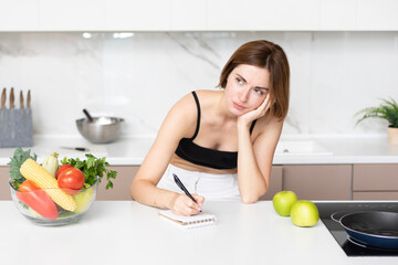 Young attractive and slender woman sitting at the table in the kitchen and writing her diet plan in notebook. A lot of fruits and vegetables which on the table also included in her ration