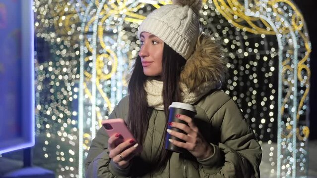 Young woman using her phone via texting with hot coffee in hands. Winter evening. Female in knitted hat with xmas lights at background. Christmas concept. 4K, UHD