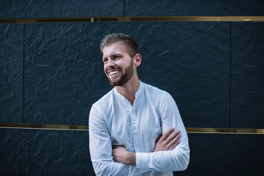 Happy adult guy standing with folded hands near dark gray wall