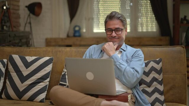 Happy middle aged man sitting on couch at home working with laptop computer, smiling.