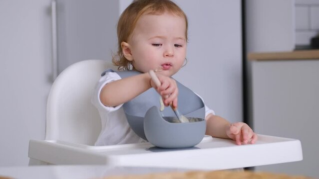 4K video Cute baby learning self-feeding. Infant child wearing bib siting in highchair eating holding a spoon by himself. Developmental milestones.