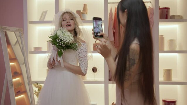 Bridesmaid takes mobile pictures of her friend wearing wedding dress. Girls having fun in wardrobe.