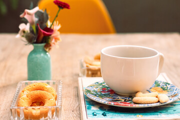 Xicara com pirex encima de livro, flores, biscoitos e café da manha na mesa com cadeira