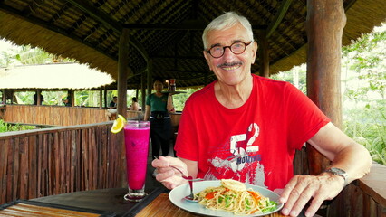 aktiver Senior genießt fröhlich sein Essen in einem Restaurant auf Stelzen über dem Dschungel...