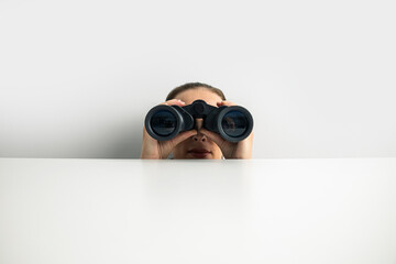 Young woman looking through binoculars peeking out from behind white cardboard on white background